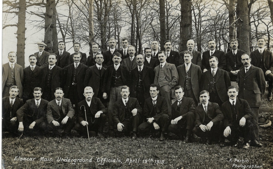 A black-and-white photo of dozens of men in suits and ties posing outdoors among bare trees. The caption reads, "Elsecar Main Underground Officials, April 19th 1919.